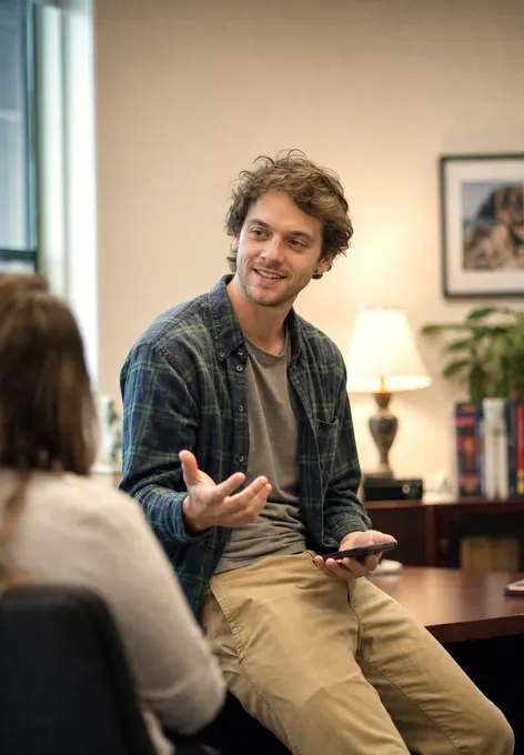 An academic advisor sitting on the edge of his desk, mid-conversation with a student, phone in one hand
