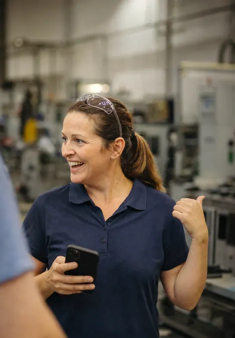 An operations lead in a workshop full of machinery, mid-conversation, grinning and pointing back over her shoulder