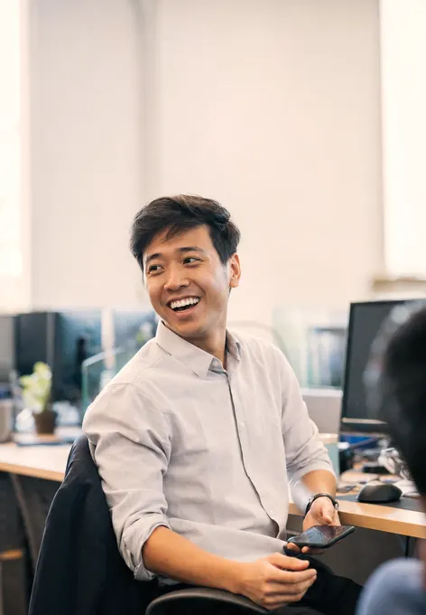 A finance analyst turning from two monitors to laugh with a colleague, phone in hand