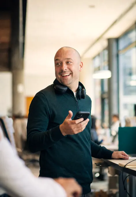 A customer-service lead at a stand-up desk turning to answer someone, headset round his neck, phone raised