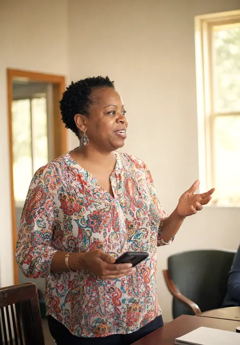 An executive director standing at the head of a small meeting table mid-sentence, smiling, phone in hand